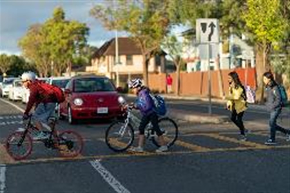 Four children with bookbags in a crosswalk, with cars stopped for them to cross. One child rides a bike, one walks a bike, and two are walking. 