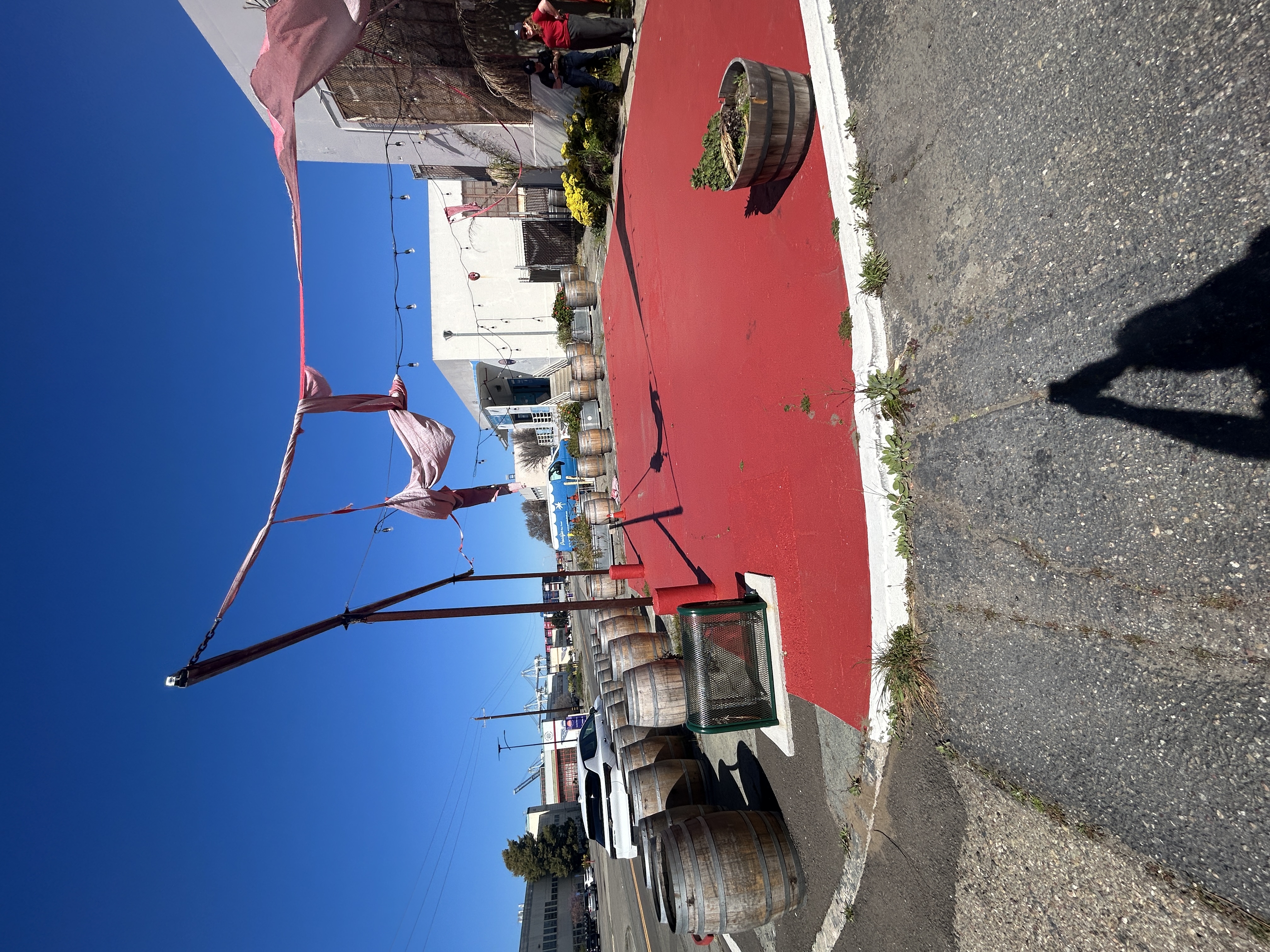 Exterior view of concrete/metal posts supporting tattered red shades, outdoor string-lights, wooden wine barrels with metal rings