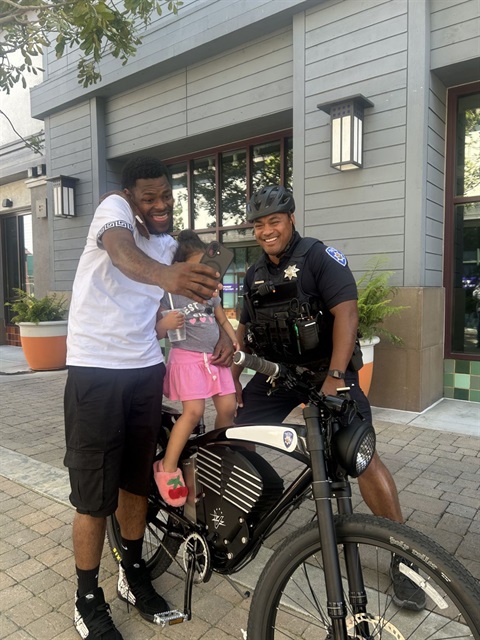 Picture an APD bike patrol officer posing for a selfie with a community member and their child on a police bike. 