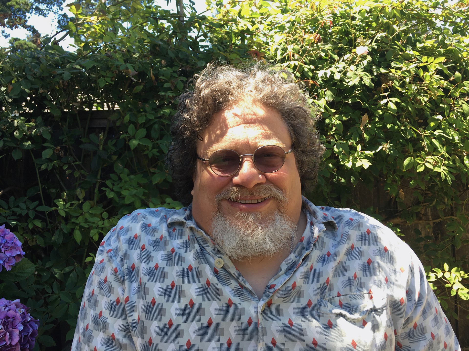 Co-director Jim LeBrecht smiles in front of green plants, sun shining on his face.